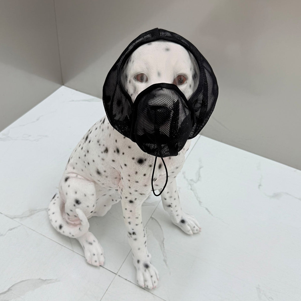 A black mesh dog muzzle and neck collar set worn by a white Dalmatian dog sitting on a light marble surface.