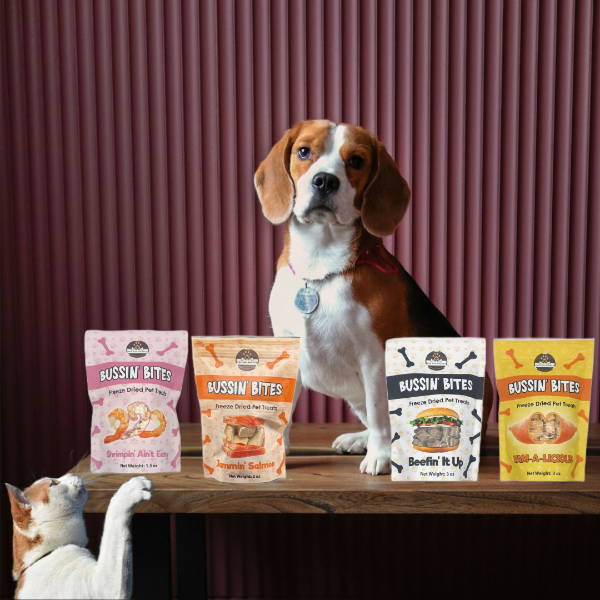 Dog and cat sitting behind a table with Bussin Bites pet food packages on a wooden surface.